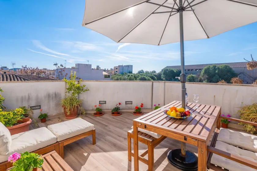 A sunny rooftop terrace has a wooden dining table beneath a large umbrella.