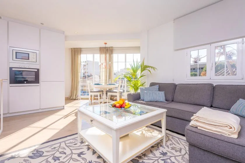 A bright living room with a gray sectional, glass coffee table, and dining area by the windows.