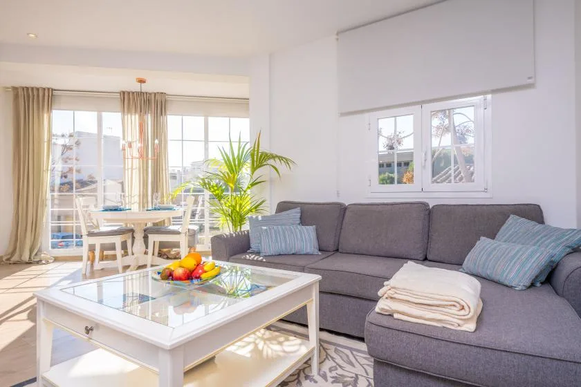 A bright living room with gray seating, a glass coffee table, and a dining area by the windows.