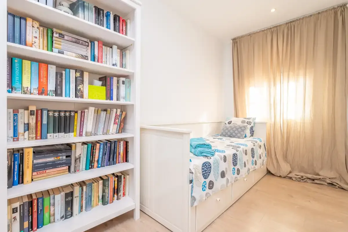 A bright bedroom with a single bed beside a tall bookshelf filled with books.