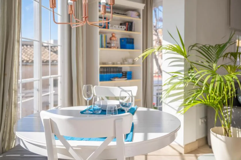 A bright dining area with a round table, white seating, and a large potted plant.