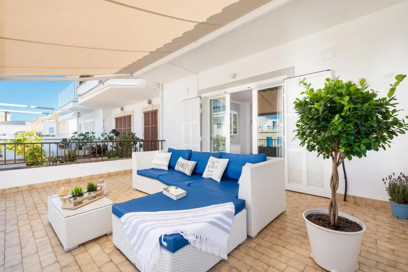A covered outdoor patio has blue seating, a low table, and potted plants.
