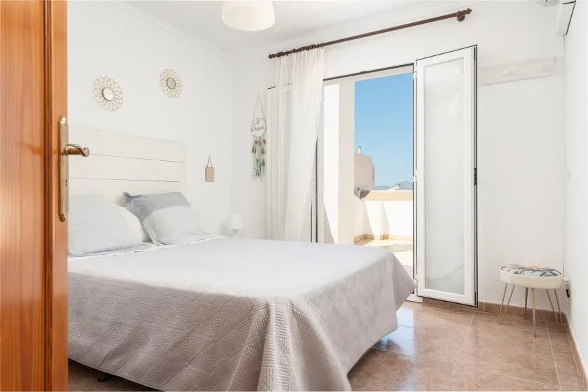A bright bedroom with a bed beside open balcony doors.