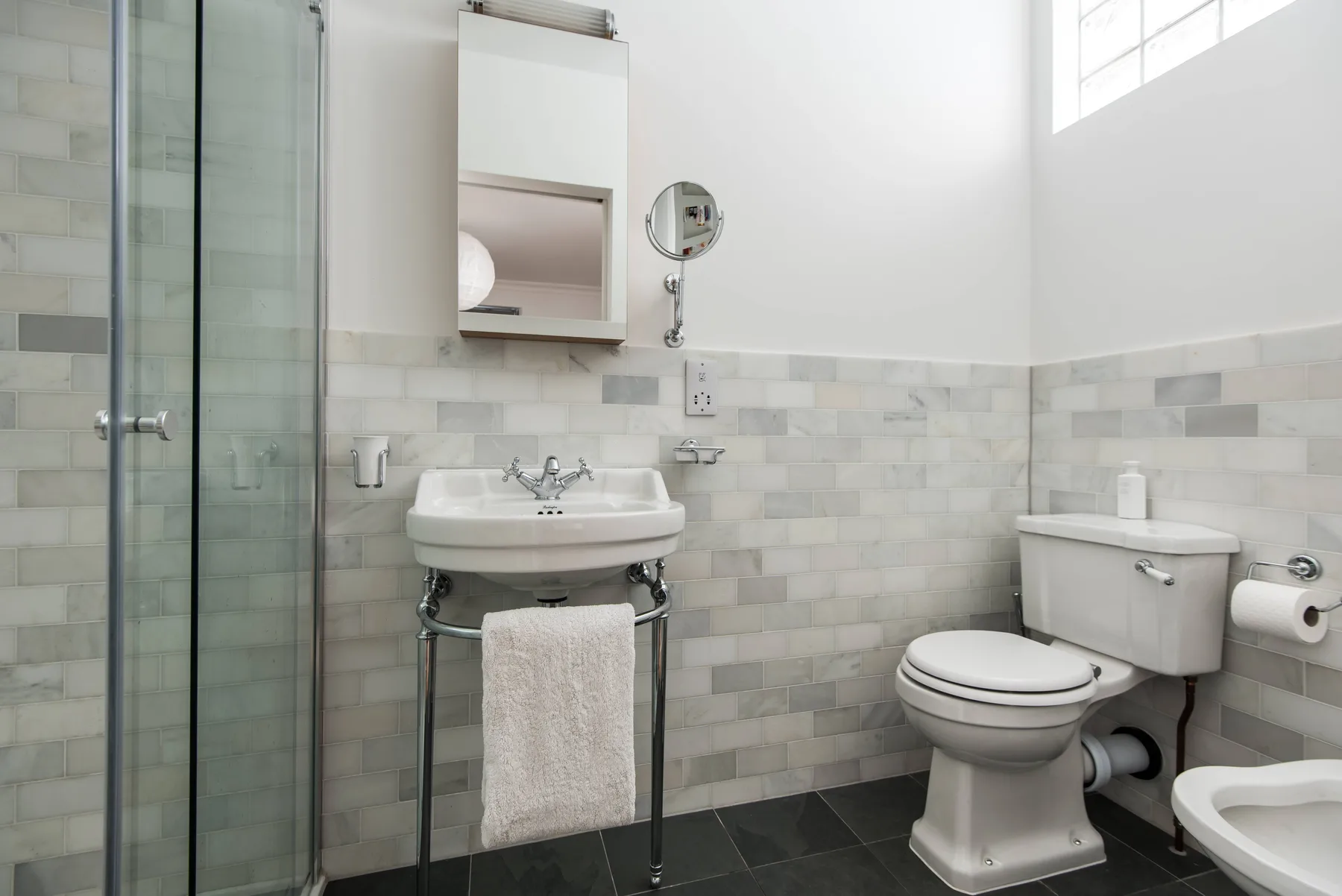 A bright bathroom with a glass shower, pedestal sink, and toilet.