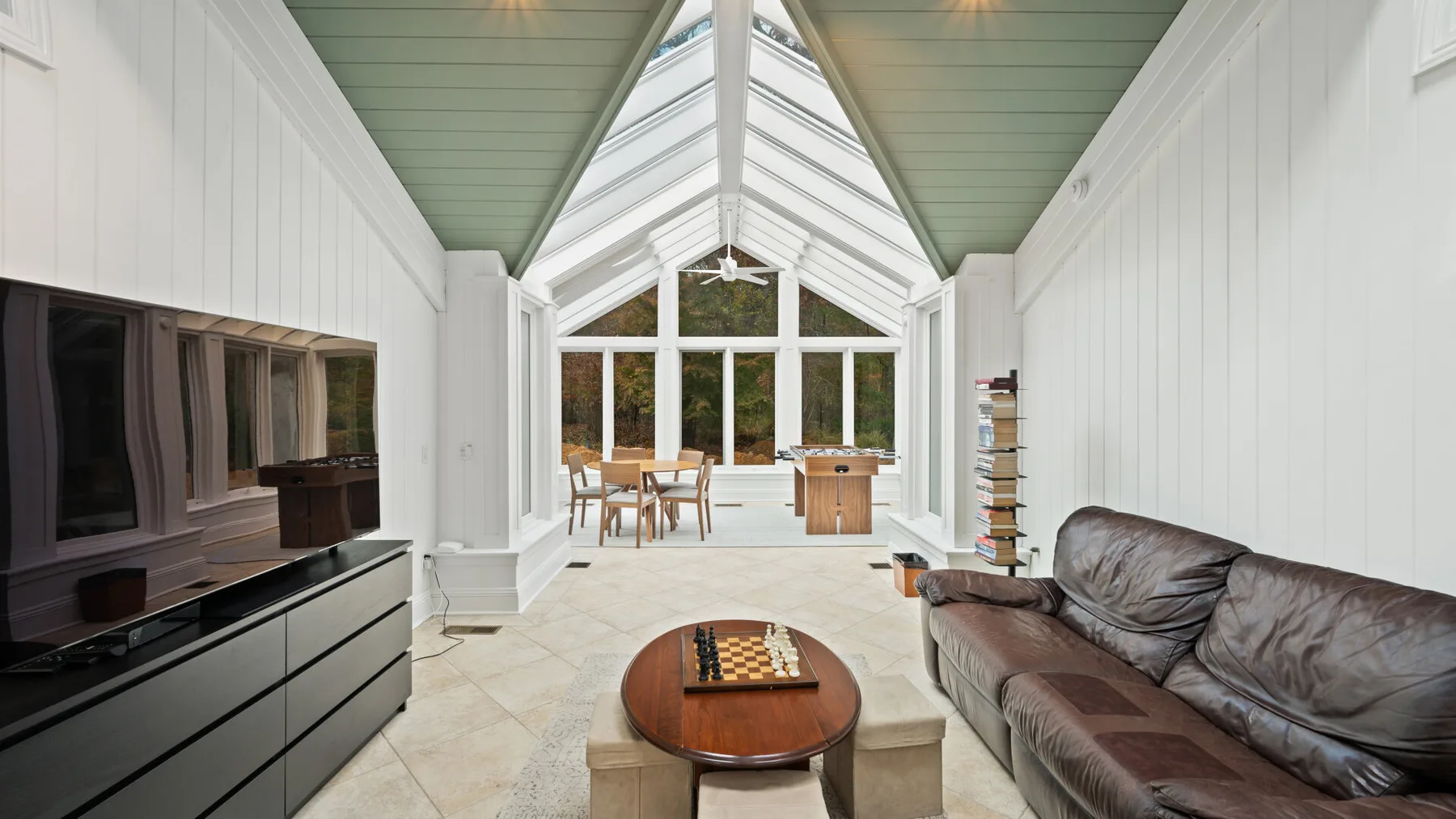 A bright sunroom with seating, a television, and a dining table beneath a glass roof.