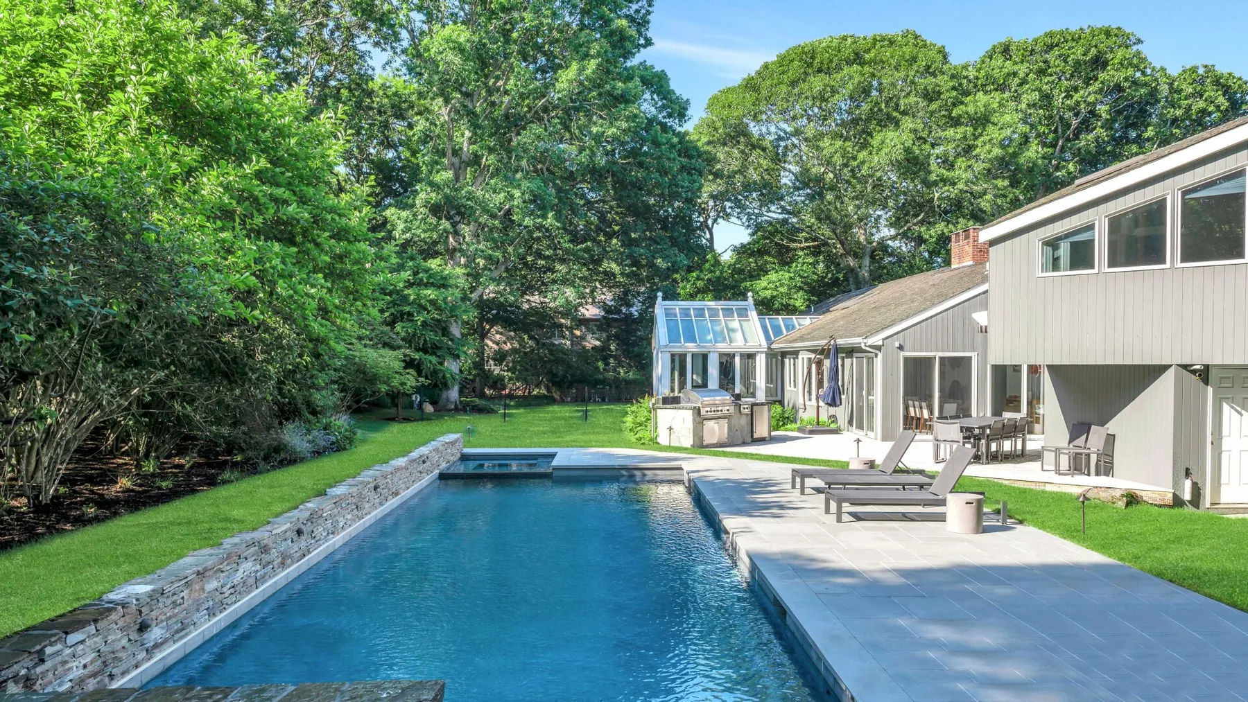 An outdoor swimming pool beside a house, with patio seating and trees.