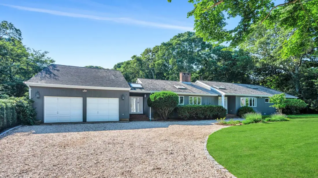 A single-story house with an attached two-car garage faces a gravel driveway and green lawn.