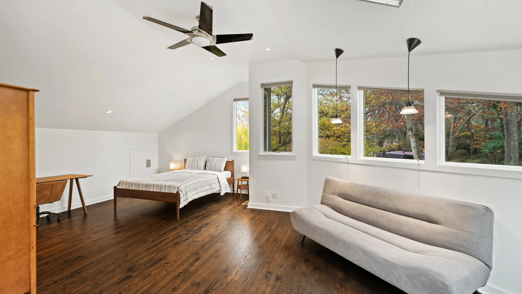 A bedroom and seating area with a bed, gray sofa, and windows overlooking trees.