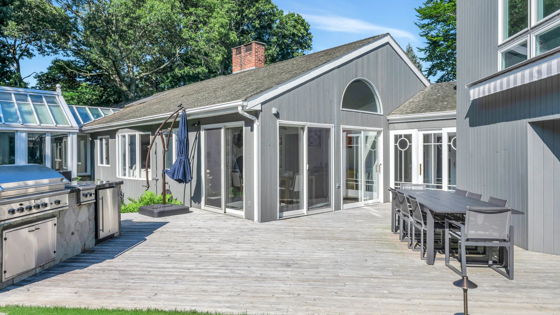 A spacious outdoor deck with a dining table and a grill beside a gray house.