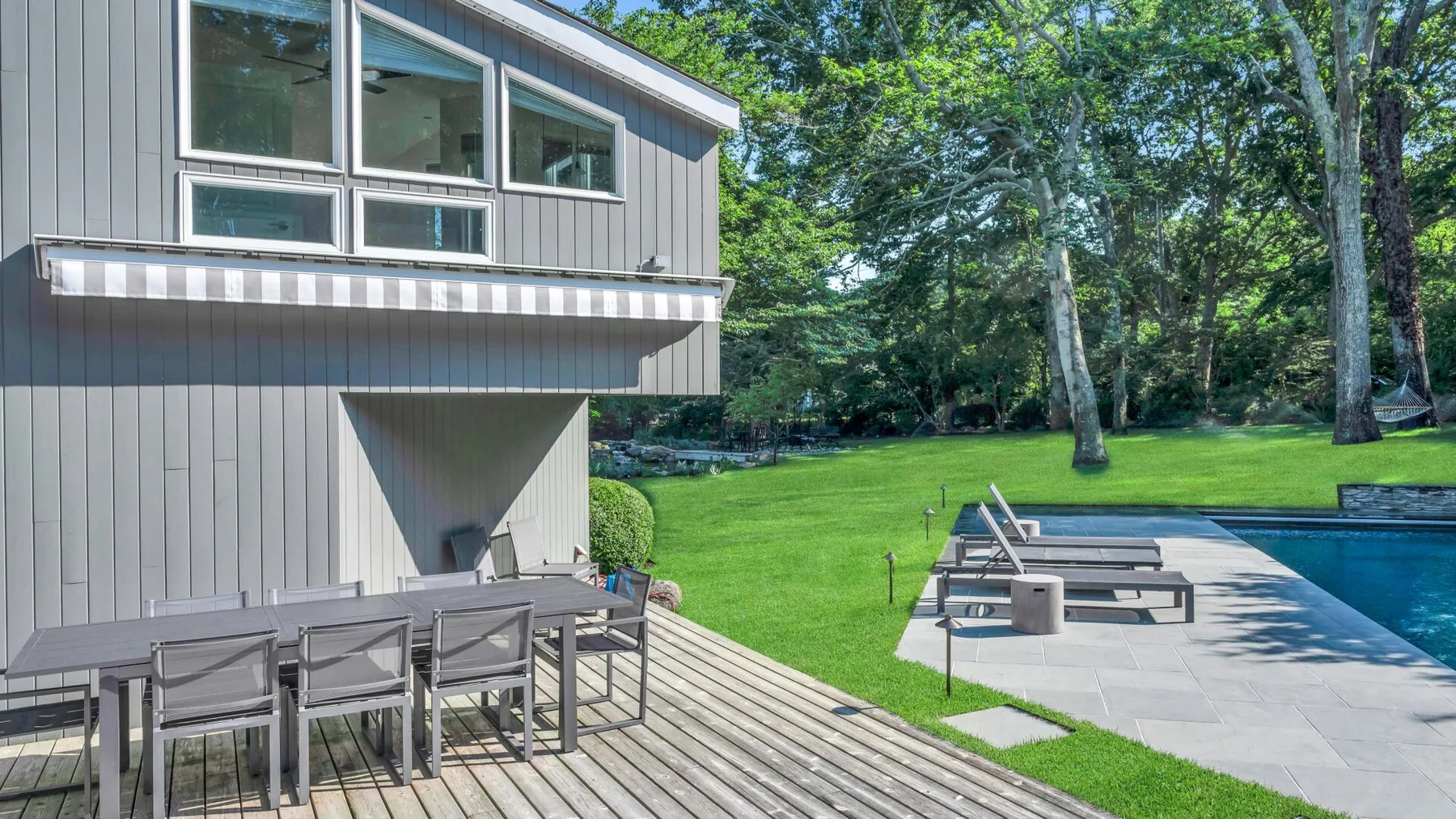 A modern house opens onto a wooden deck beside a lawn and swimming pool.