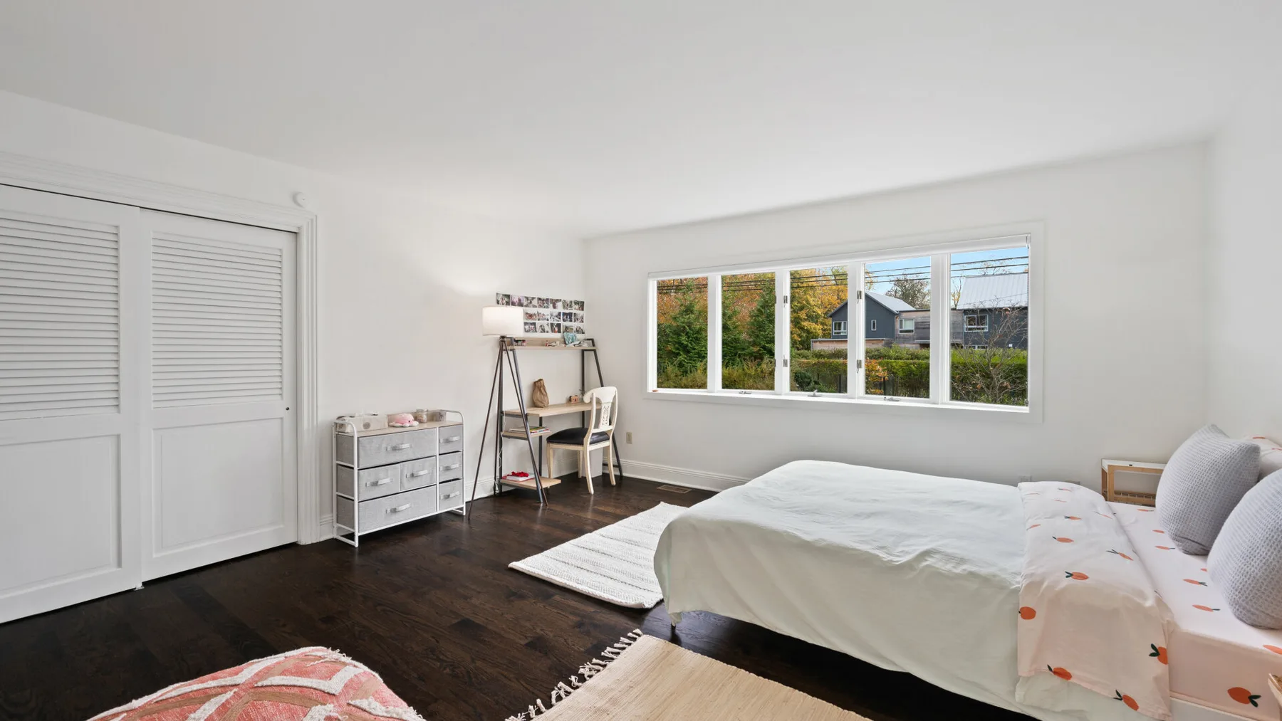 A bright bedroom with a bed, dark flooring, and a wide window overlooking greenery.