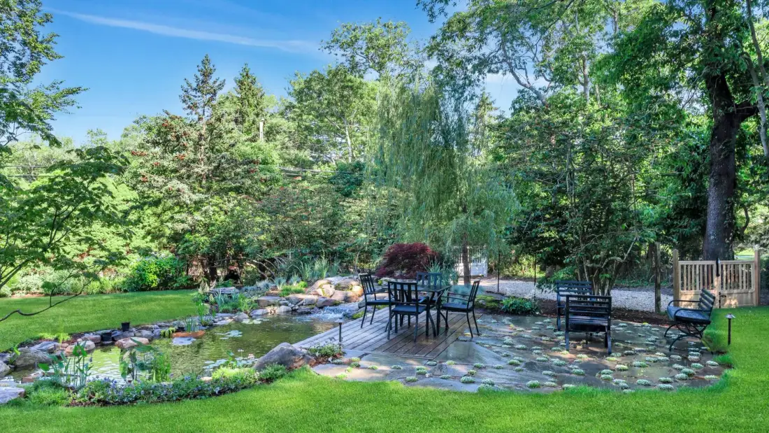 Un patio de jardín con asientos al aire libre da a un estanque rodeado de árboles.