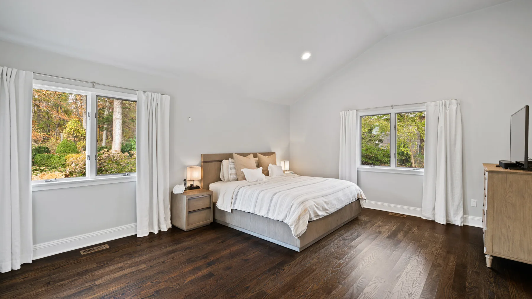 A bright bedroom with a bed between two windows and dark wood flooring.