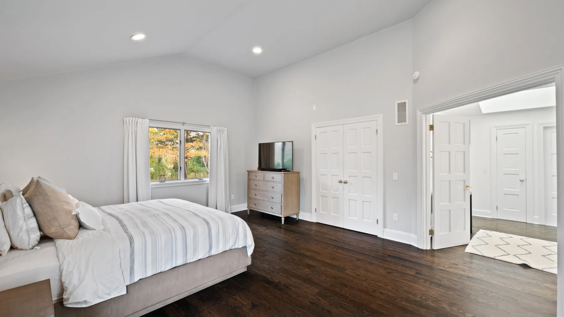 A bright bedroom with a bed, a window, and dark wood flooring.