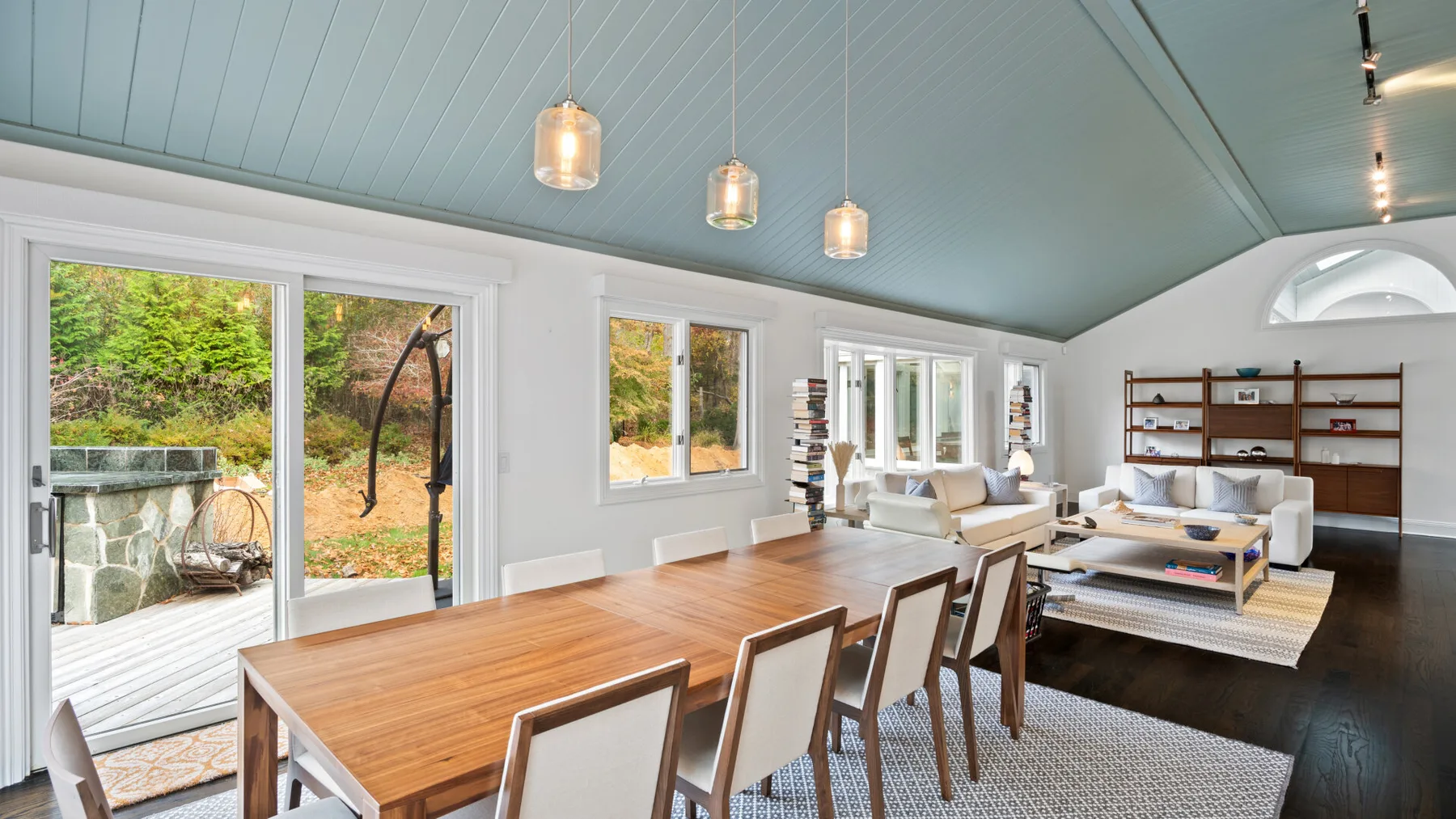 A dining area opens onto a living room, with large windows overlooking greenery.