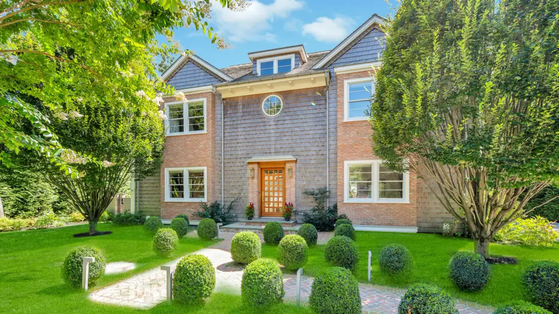 A large brick-and-shingle house stands behind a lawn and neatly trimmed shrubs.