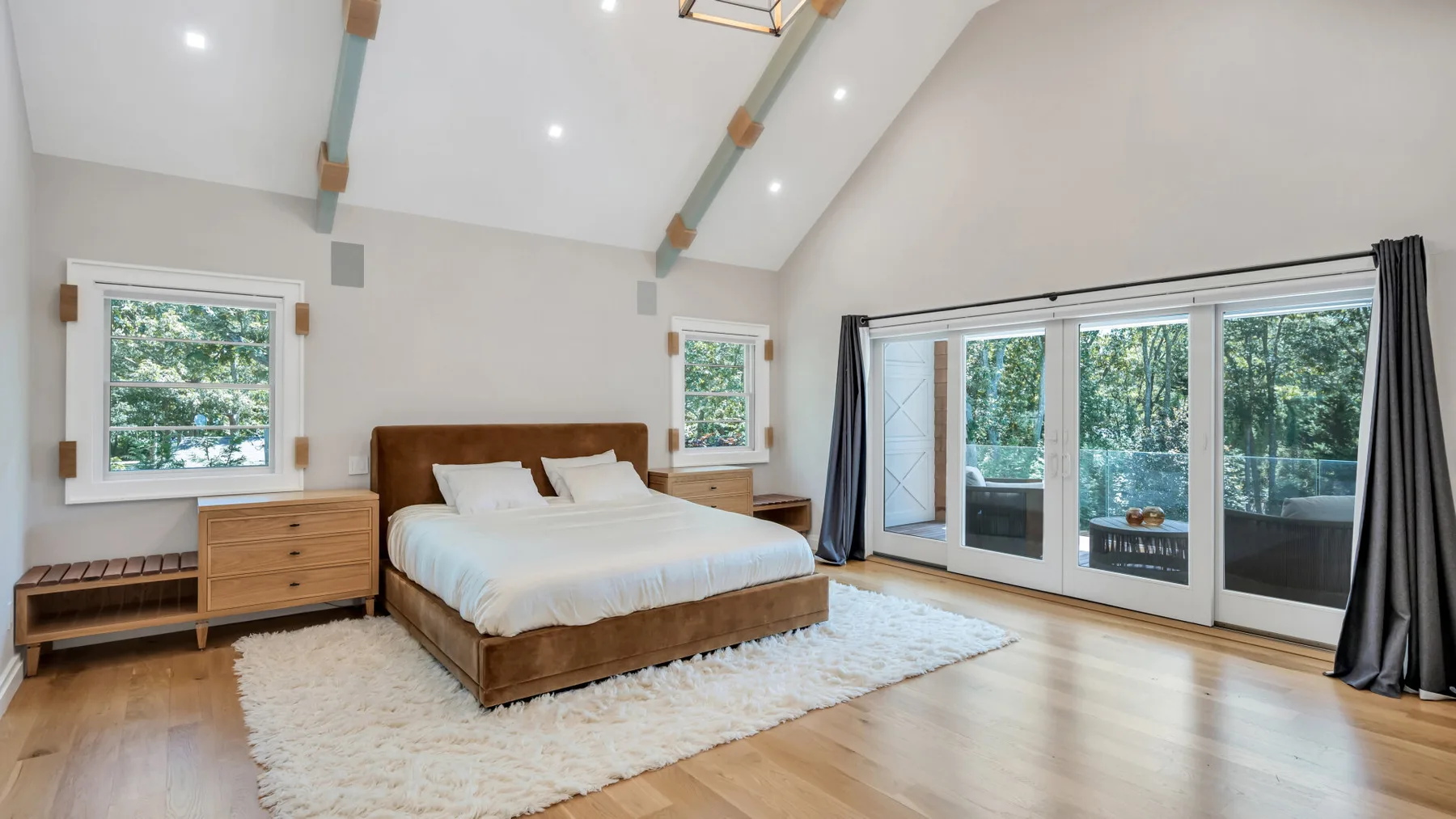 A bright bedroom has a wooden bed, light bedding, and large glass doors.