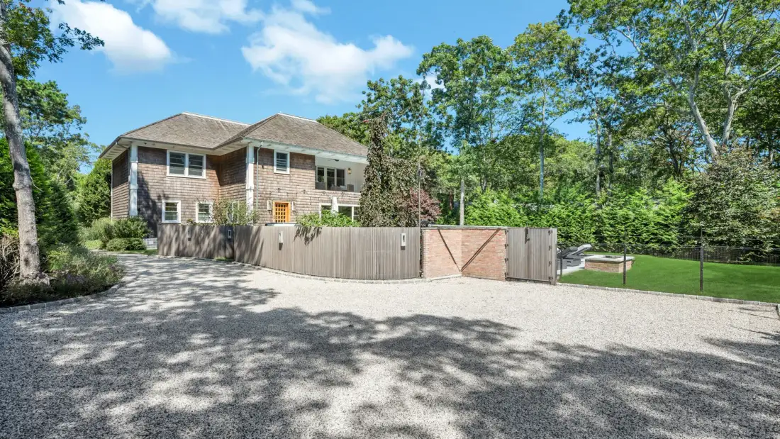 A two-story house stands behind a wooden gate, with trees around the driveway.