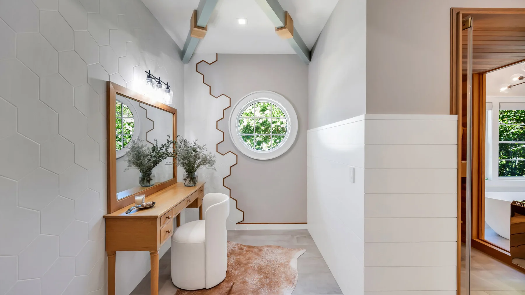 A bright dressing area features a wooden vanity, round window, and patterned rug.