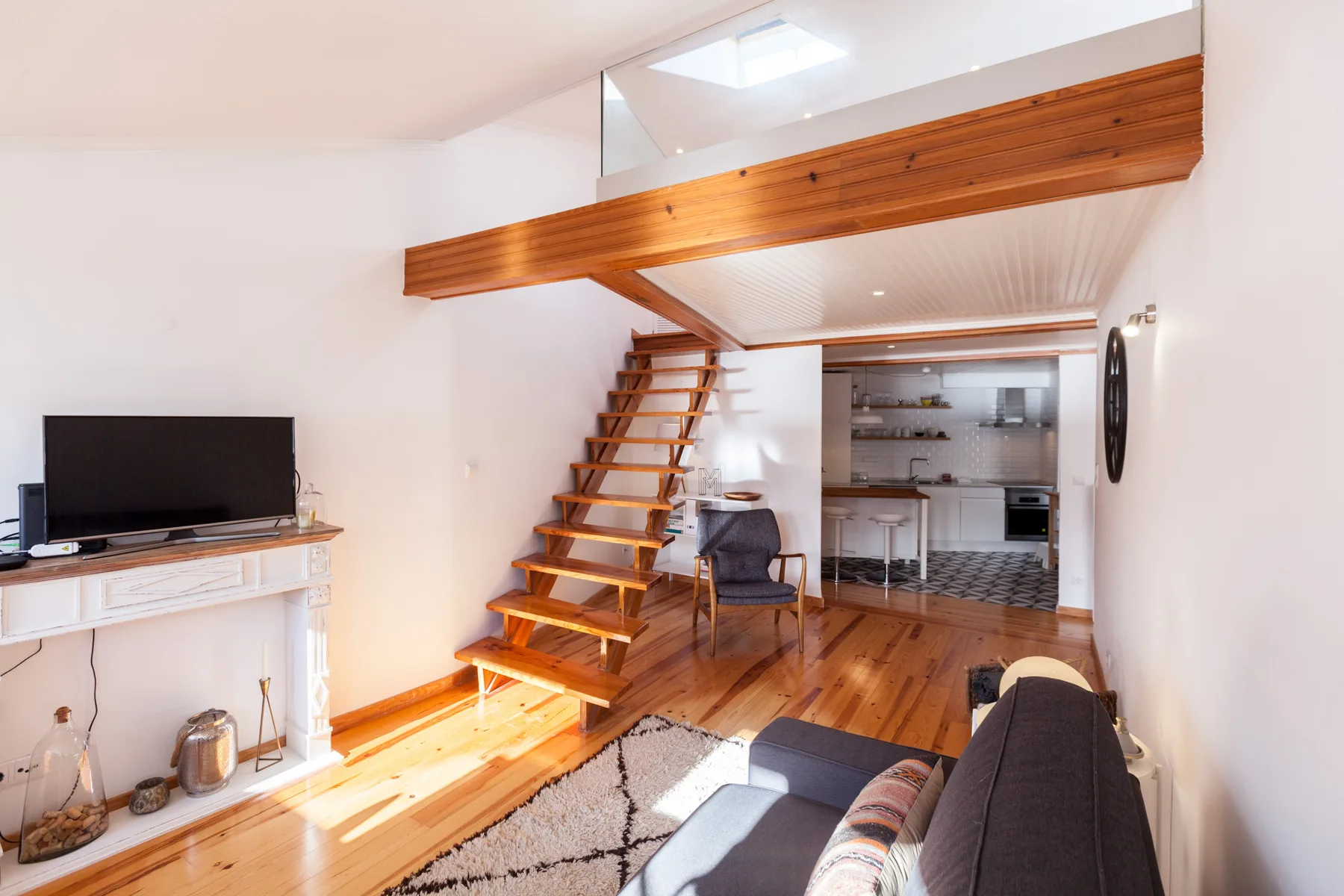 A living room with a staircase leading to a wooden mezzanine and a kitchen beyond.