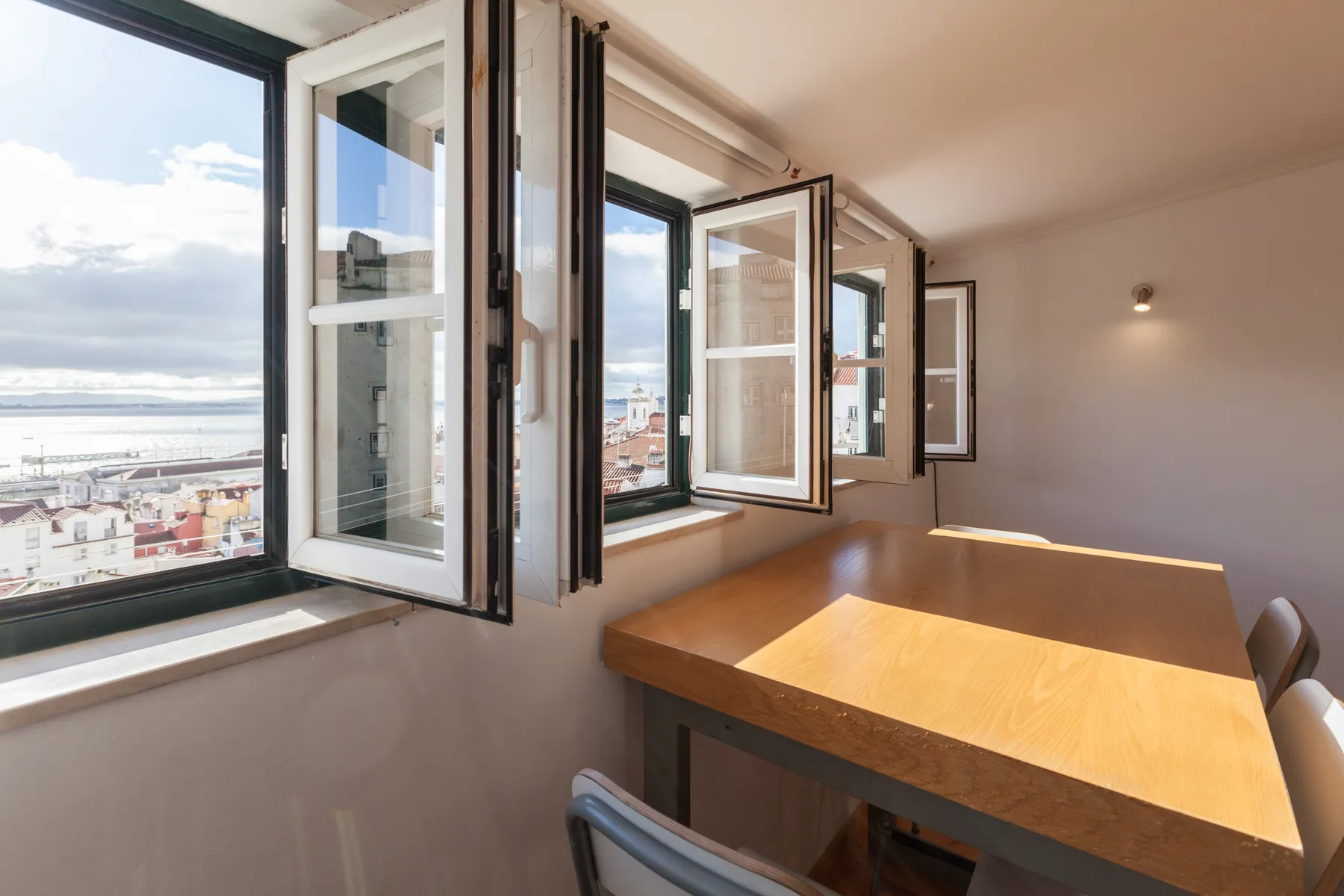 A bright dining area with a wooden table beside open windows overlooking the waterfront.