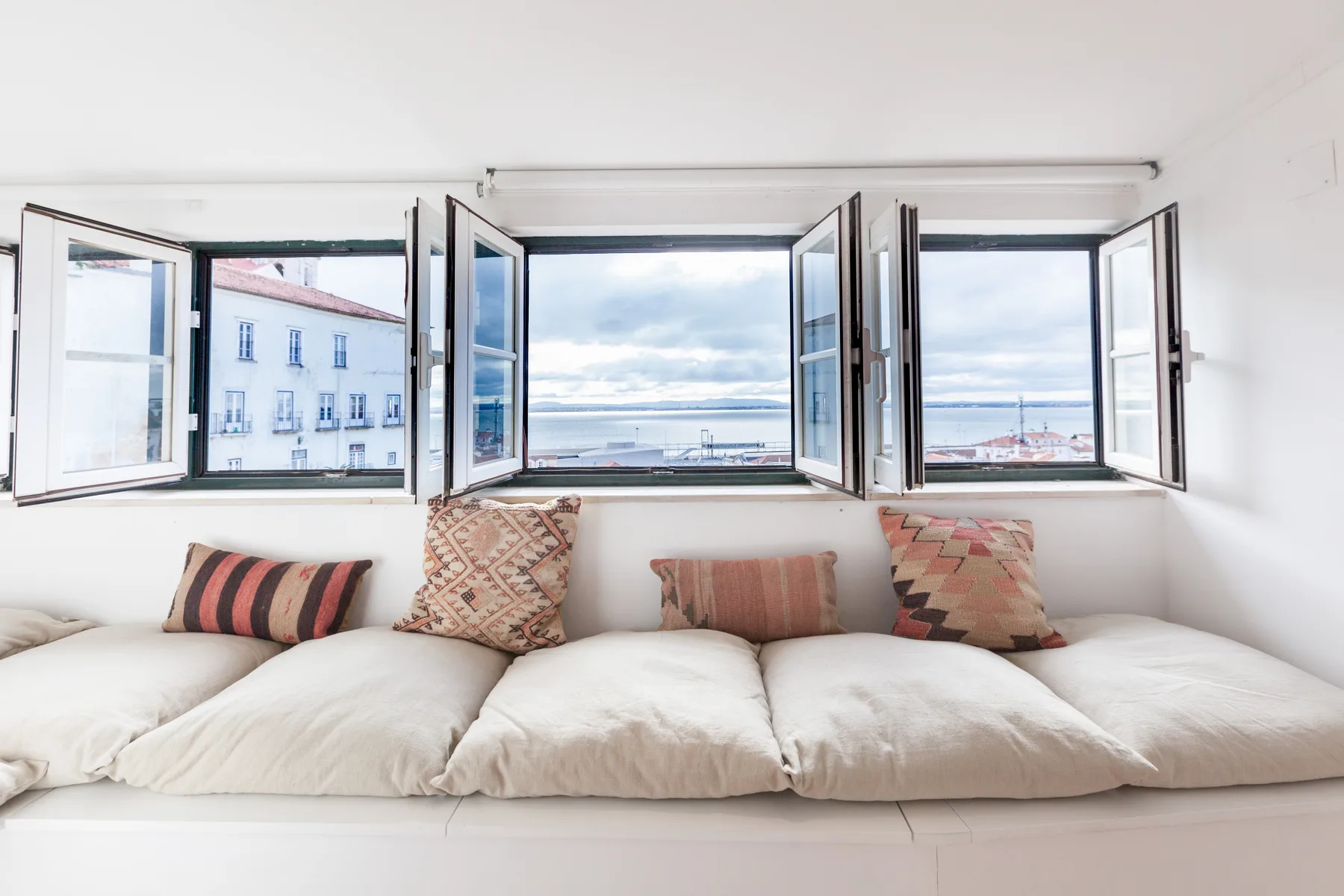 A bright sitting area with floor cushions beneath open windows overlooking the water.
