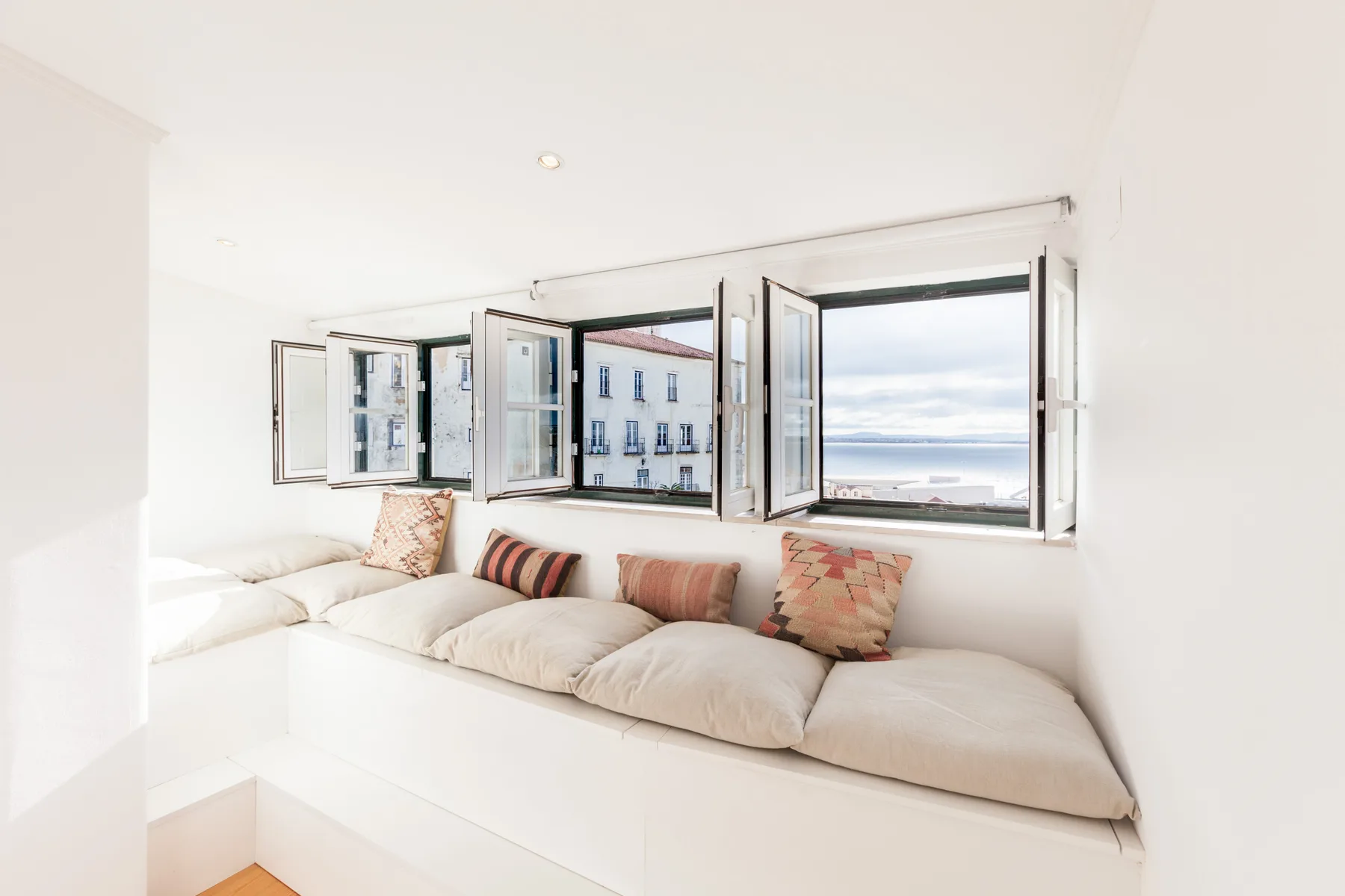A bright living area with cushioned seating beside open windows overlooking the water.
