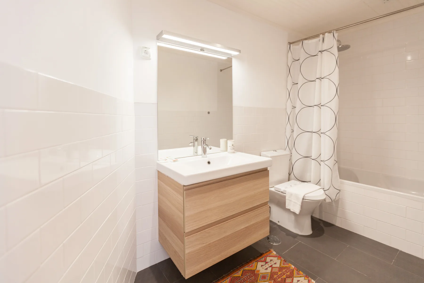 A bright bathroom has a wood-front vanity, mirror, and patterned shower curtain.