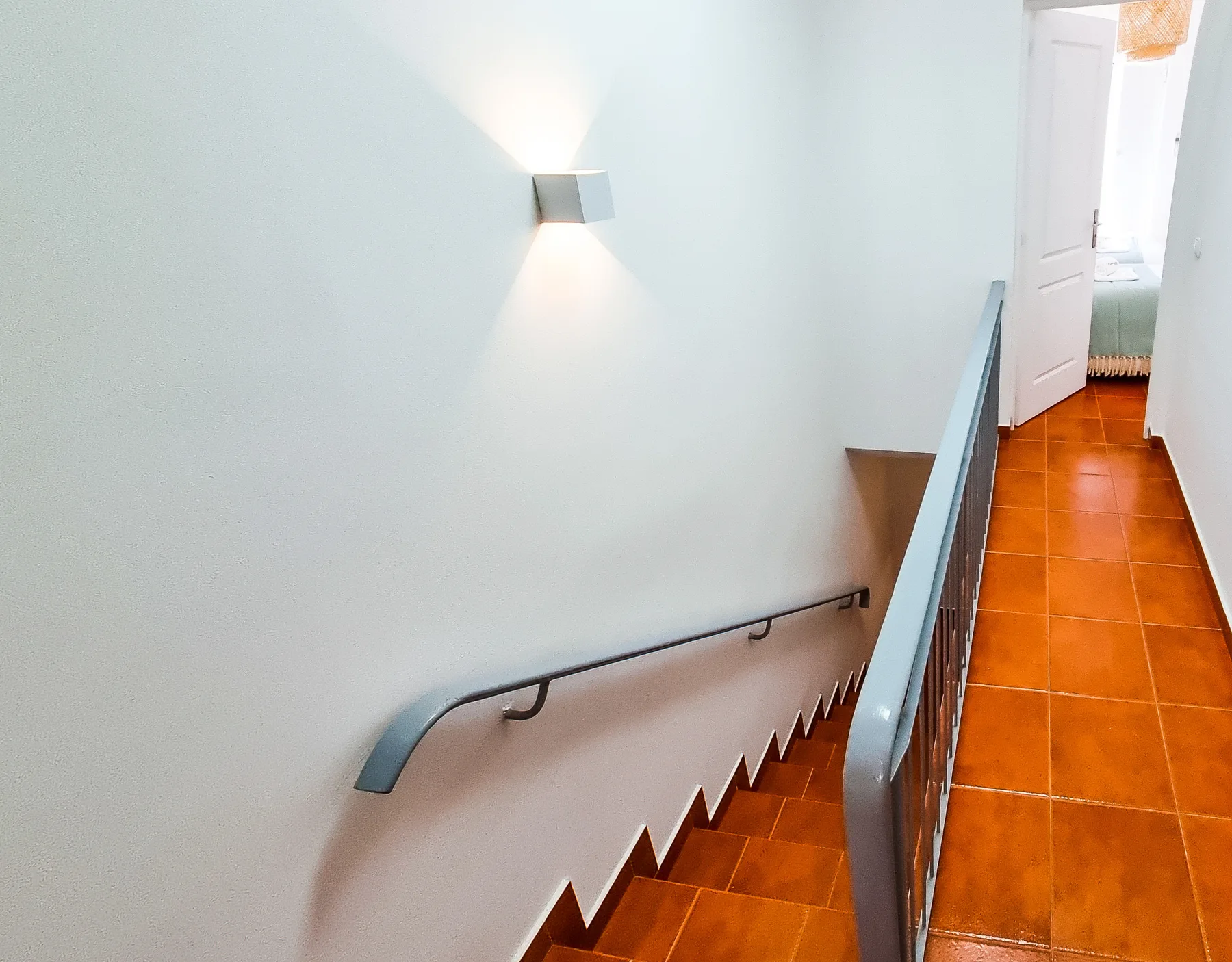A stairway with a handrail leads to a hallway with terracotta-colored floor tiles.