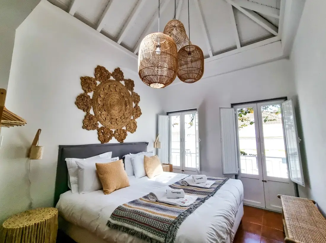 A bright bedroom with a large bed, woven pendant lights, and tall windows.