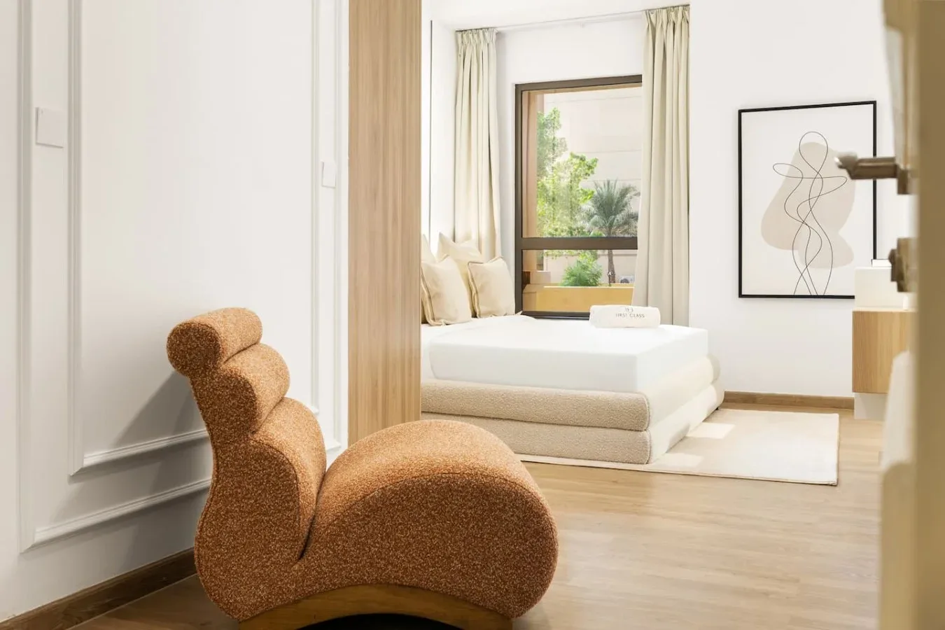 A bright bedroom with a bed, a sculptural lounge seat, and a large window.