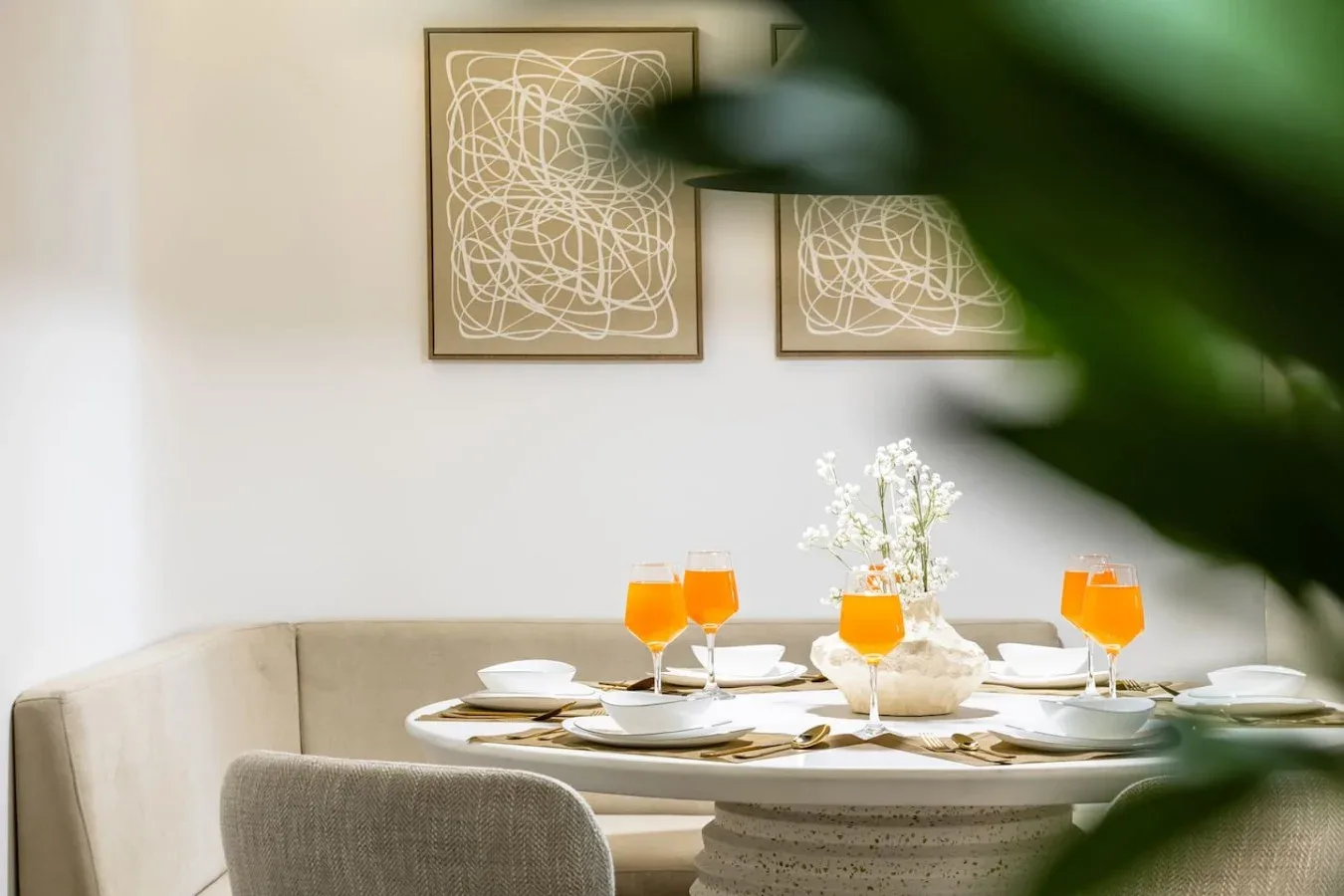 A dining nook with a set table, orange drinks, and framed abstract artwork.