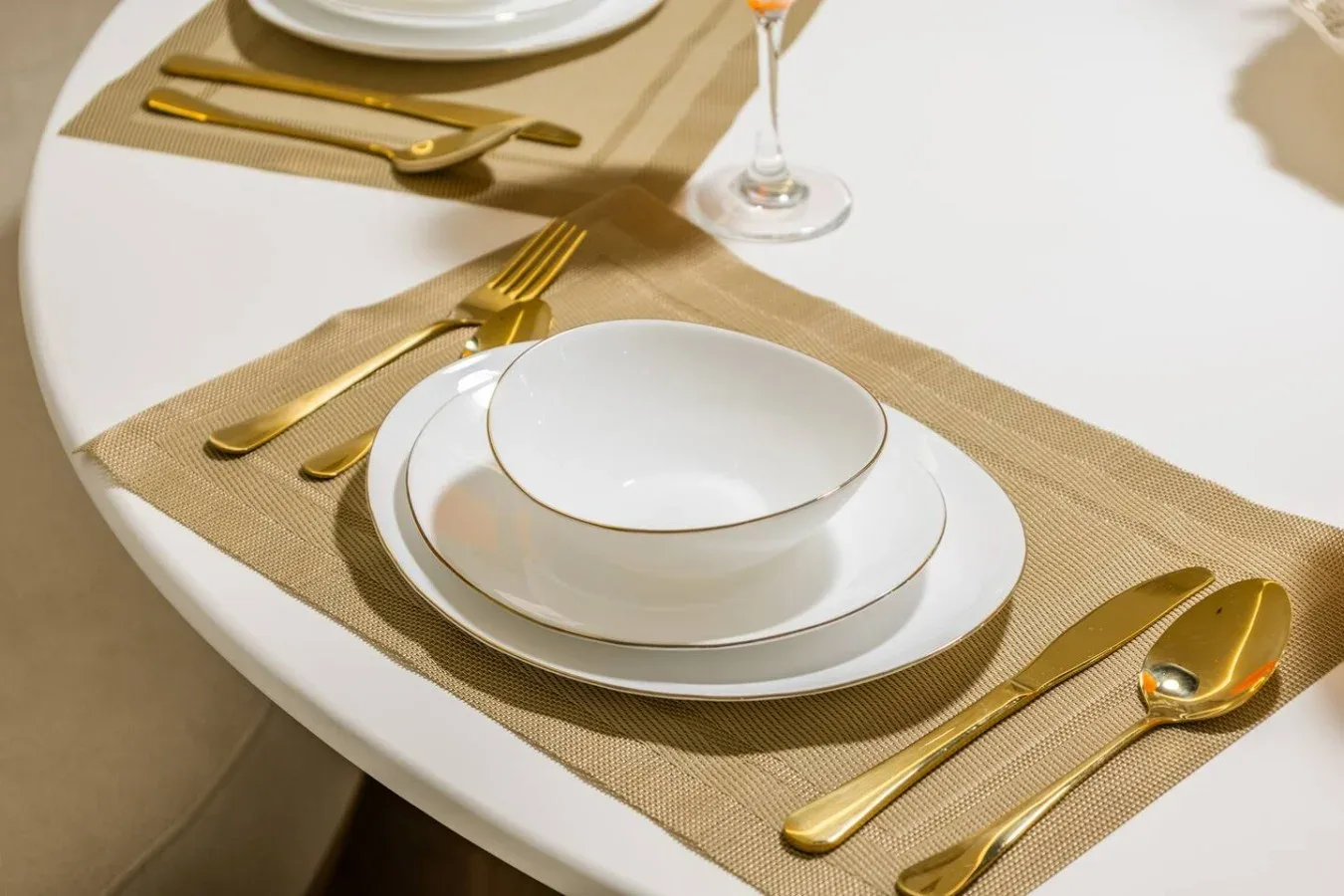 A dining table is set with white dishes, gold-colored cutlery, and woven placemats.