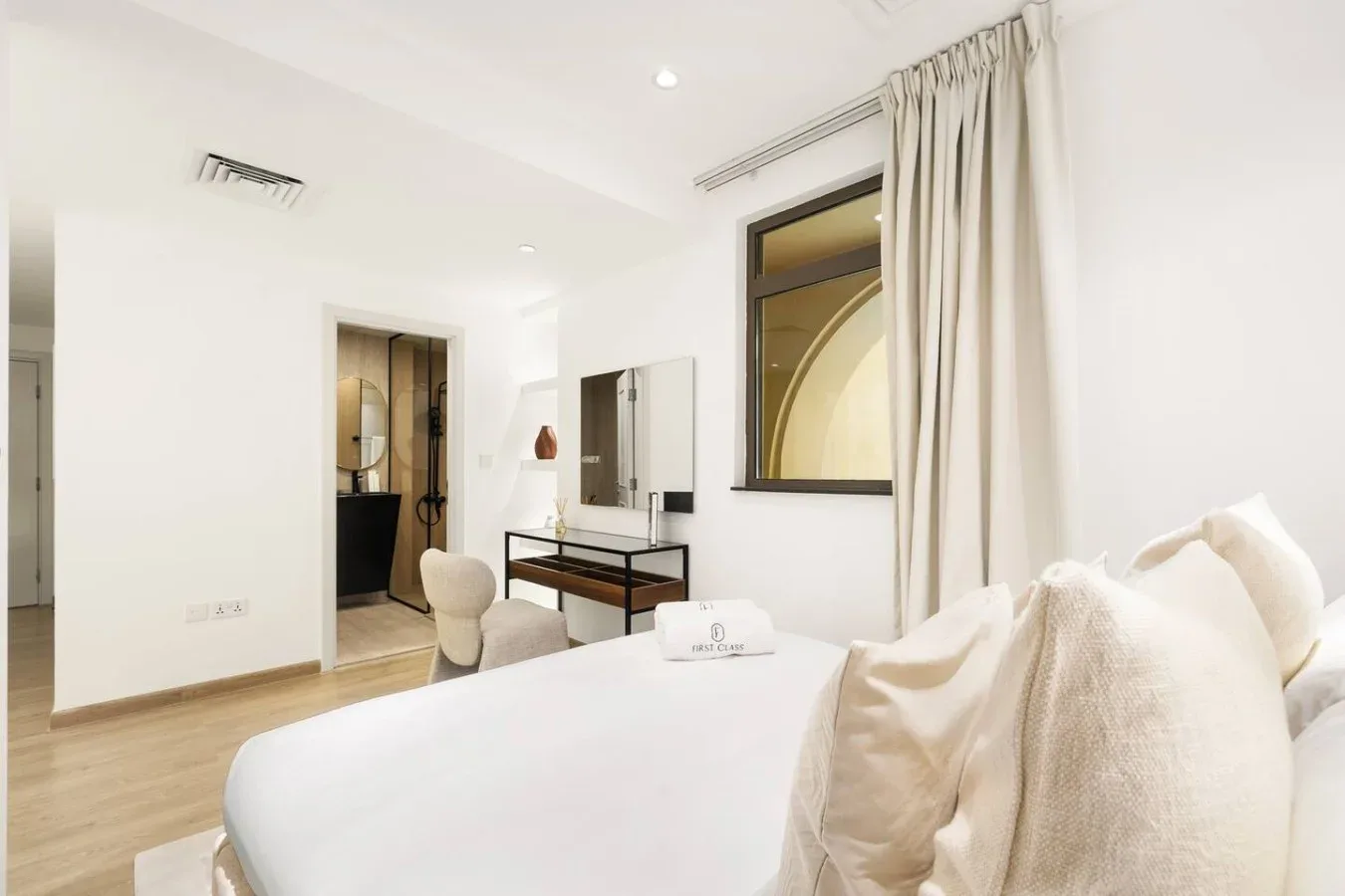 A bright bedroom with a bed, vanity, and a curtained window.