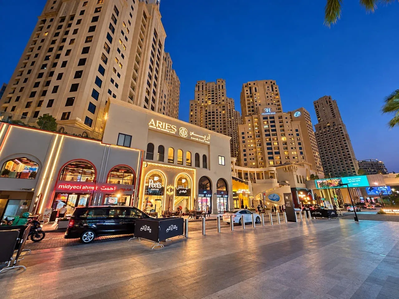 A brightly lit urban plaza with storefronts and tall buildings beneath a clear evening sky.