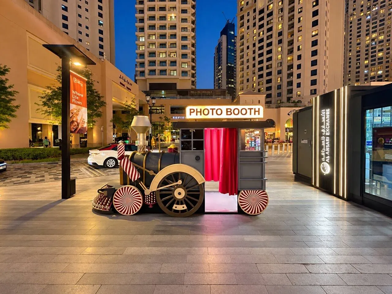 An outdoor plaza at dusk features a decorative photo booth between tall buildings.