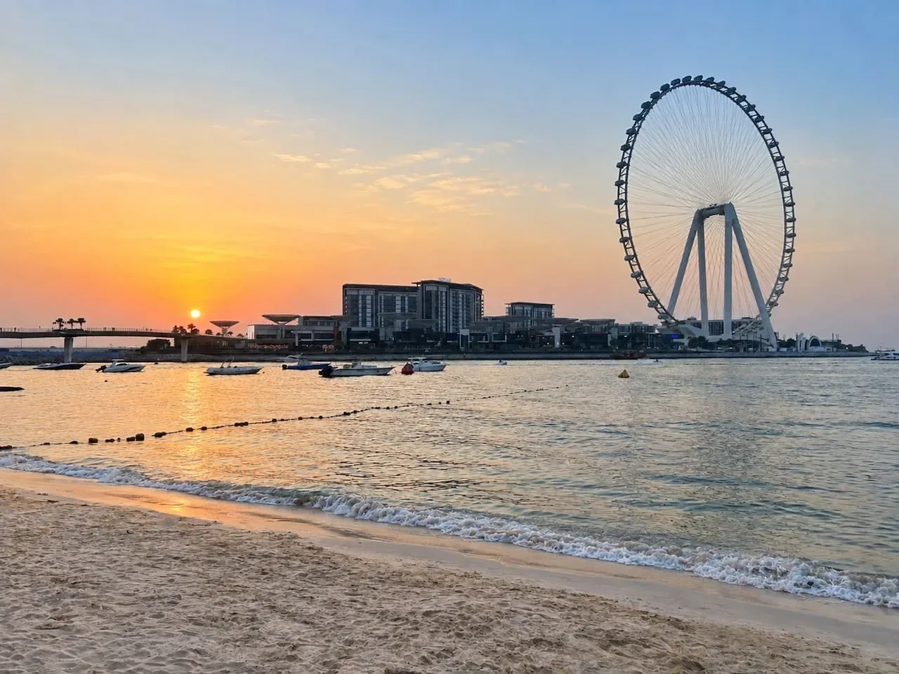 A sandy beach meets calm water beneath a sunset sky, with a large observation wheel in the distance.