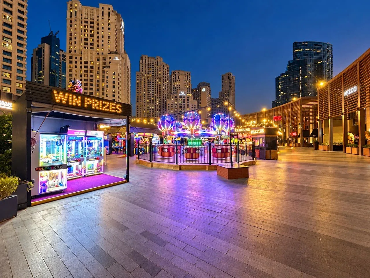A brightly lit outdoor amusement area sits among tall city buildings at night.