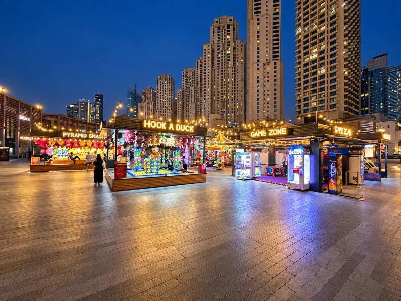 An illuminated outdoor arcade plaza with colorful game booths beneath high-rise buildings at dusk.