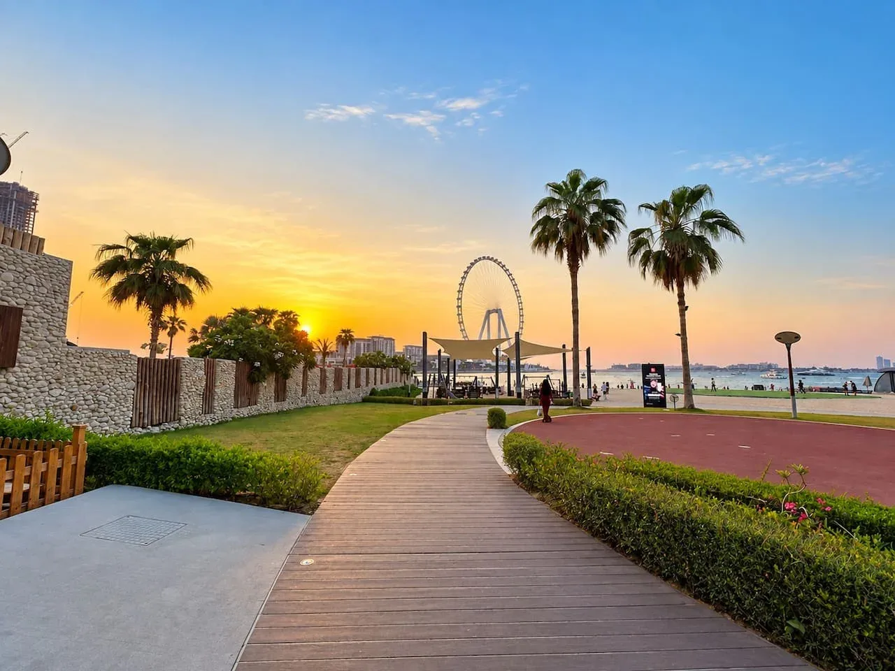 A waterfront promenade curves past landscaped greenery toward a large observation wheel at sunset.