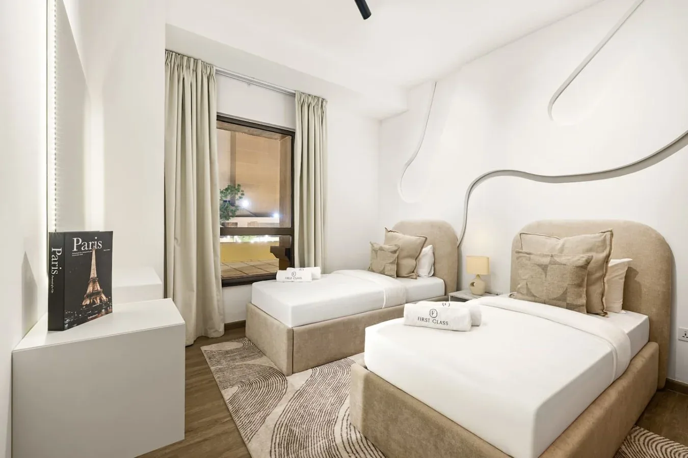 A bright bedroom with two beds, neutral bedding, and a large window.