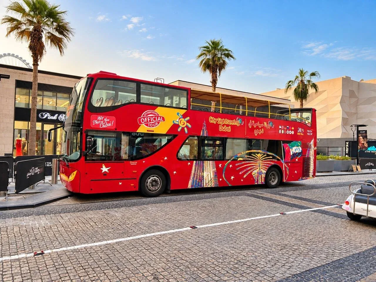 A red sightseeing bus is parked along a paved street beside modern buildings and palm trees.
