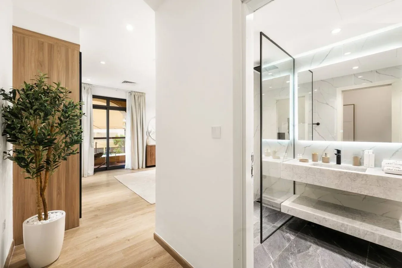 A bright bedroom area opens beside a bathroom vanity and glass shower.