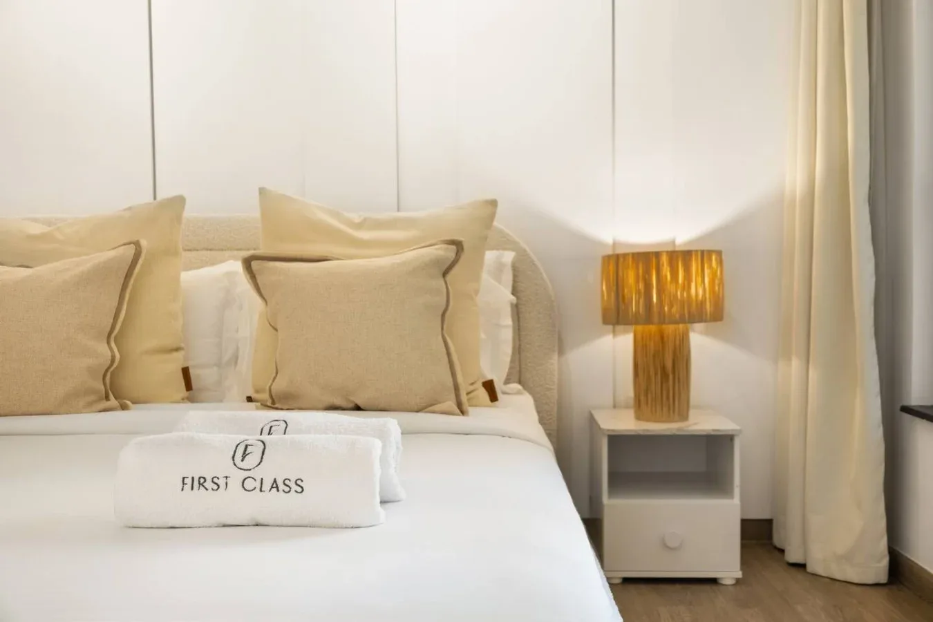 A neatly made bed with beige pillows sits beside a glowing bedside lamp.