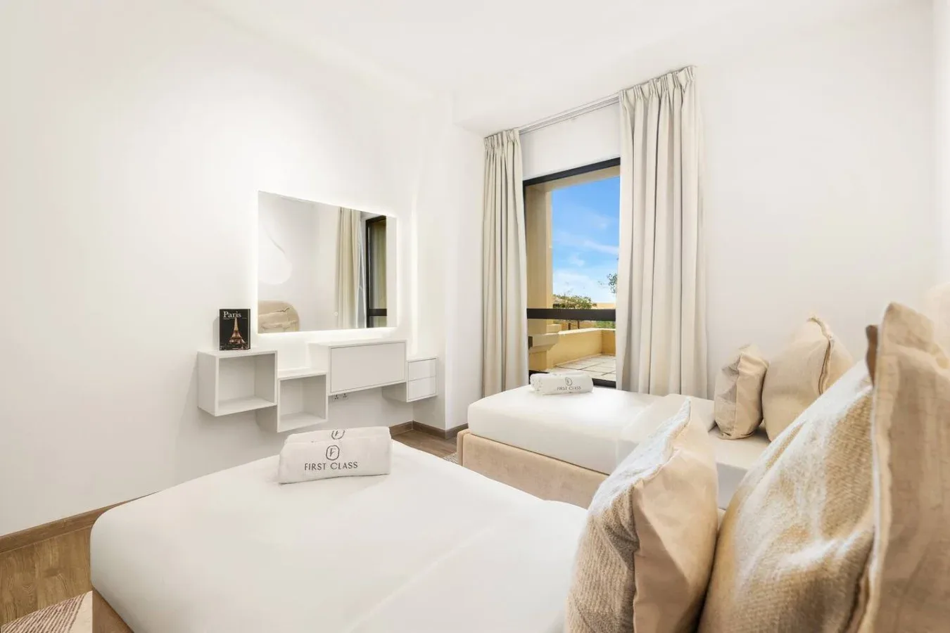 A bright bedroom with two beds, pale bedding, and a window overlooking greenery.