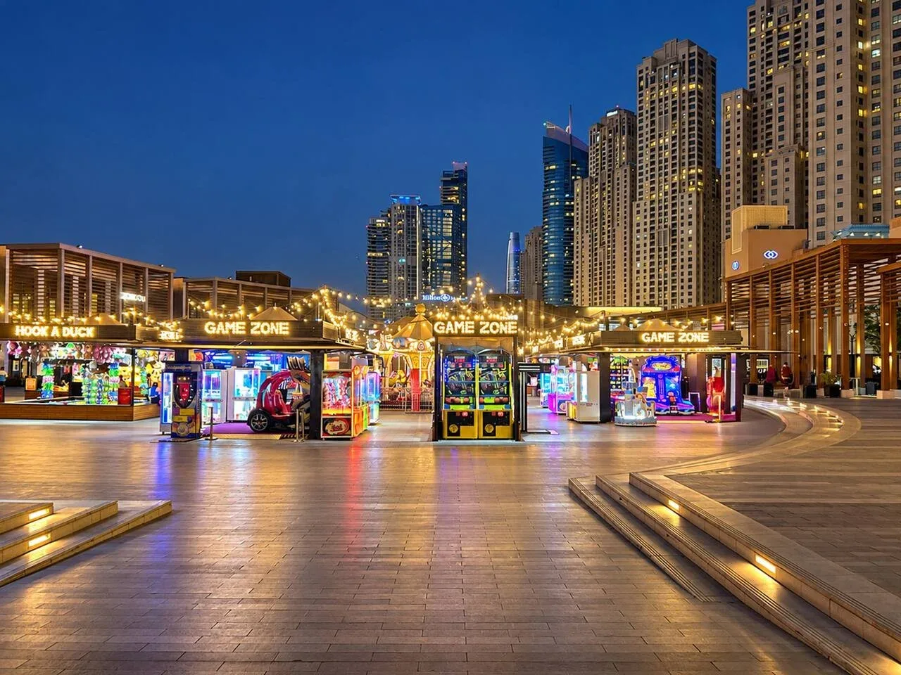 An outdoor entertainment plaza at night, with illuminated game booths and high-rise buildings.