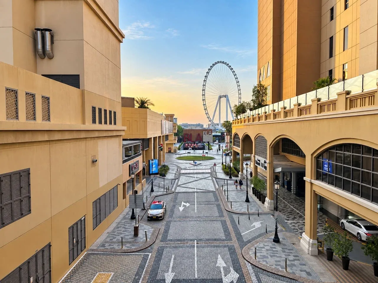 A pedestrian shopping street runs between modern buildings, with a large observation wheel in the distance.