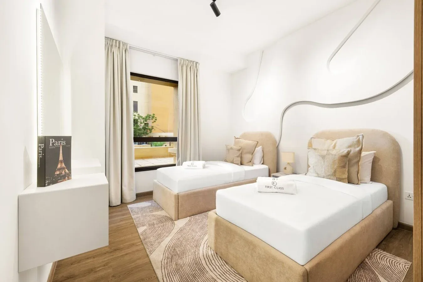 A bright bedroom with two beds, neutral bedding, and a large curtained window.