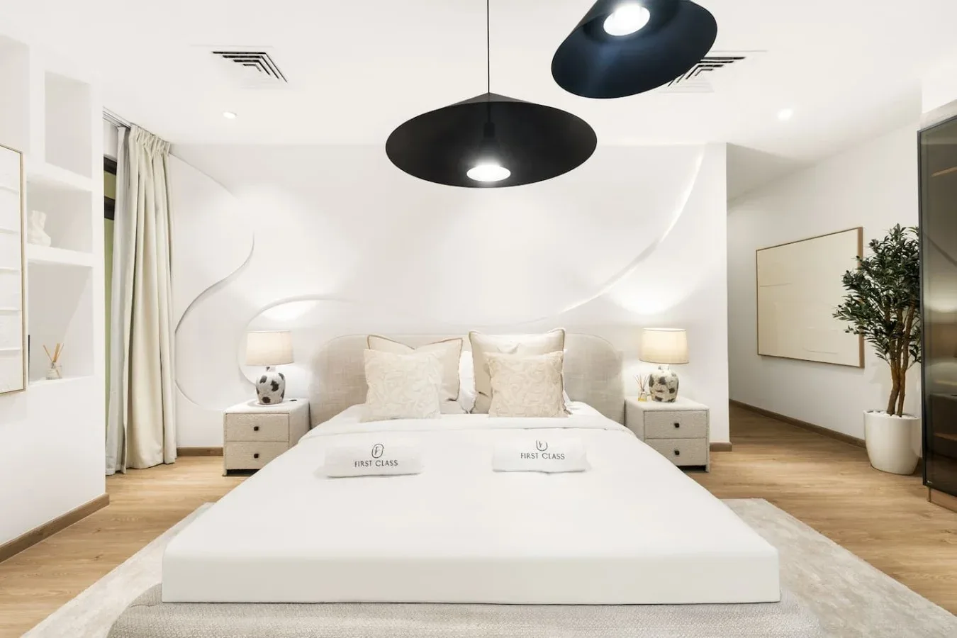A bright bedroom features a neatly made bed beneath two dark pendant lights.