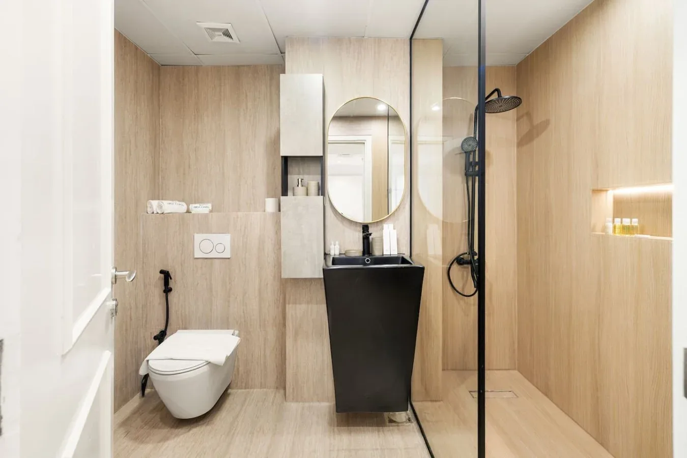 A modern bathroom with a wall-mounted toilet, dark sink, and glass shower screen.